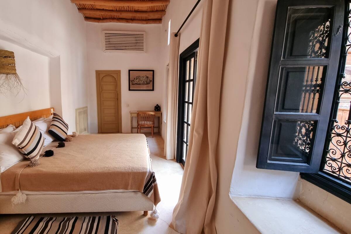 Ein Schlafzimmer mit Bett, Schreibtisch, Stuhl, Fenster mit Vorhängen und einer Tür.