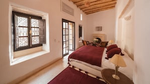 A bedroom with a wooden ceiling, a bed with a red blanket, a nightstand with a lamp, and a window with decorative iron bars. - Tagadert Lodge (Tameslouht)