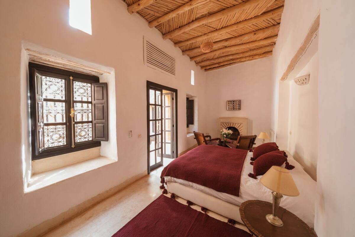 Ein Schlafzimmer mit Holzbalkendecke, einem Bett mit roter Decke, einem Nachttisch mit Lampe und einem Fenster mit dekorativen Eisenverzierungen.