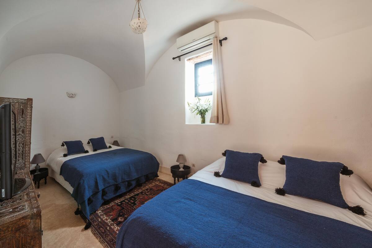 Ein Schlafzimmer mit zwei Betten, einem Fenster mit Vorhängen, einer Pendelleuchte und einem kleinen Tisch mit Lampe.