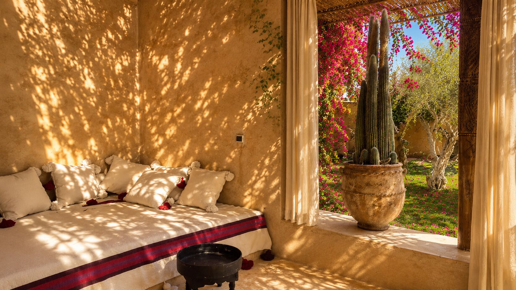 A room with a bed, a large potted cactus, and a view of greenery and flowers.