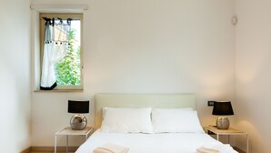 Una cama bien tendida con sábanas blancas, dos lámparas de noche y una ventana con cortinas.