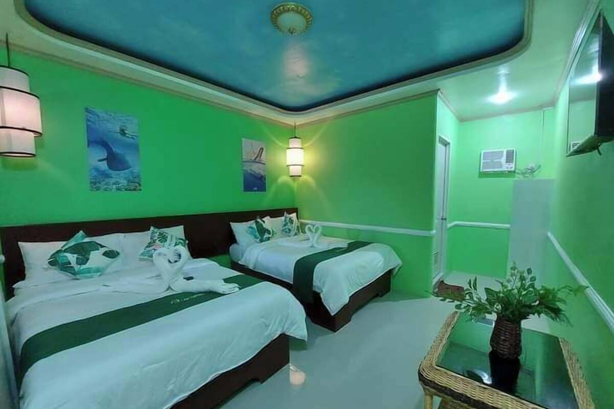 Una habitación con dos camas, paredes verdes, una lámpara en el techo y una pequeña mesa con una planta.