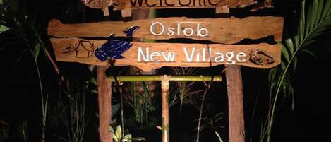 A wooden sign with 'Welcome Oslob New Village' and a bird illustration.