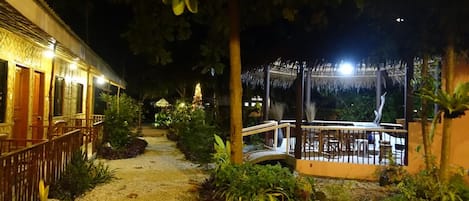 Vista nocturna de un patio de hotel con un camino, un pequeño puente y una construcción con techo de paja.