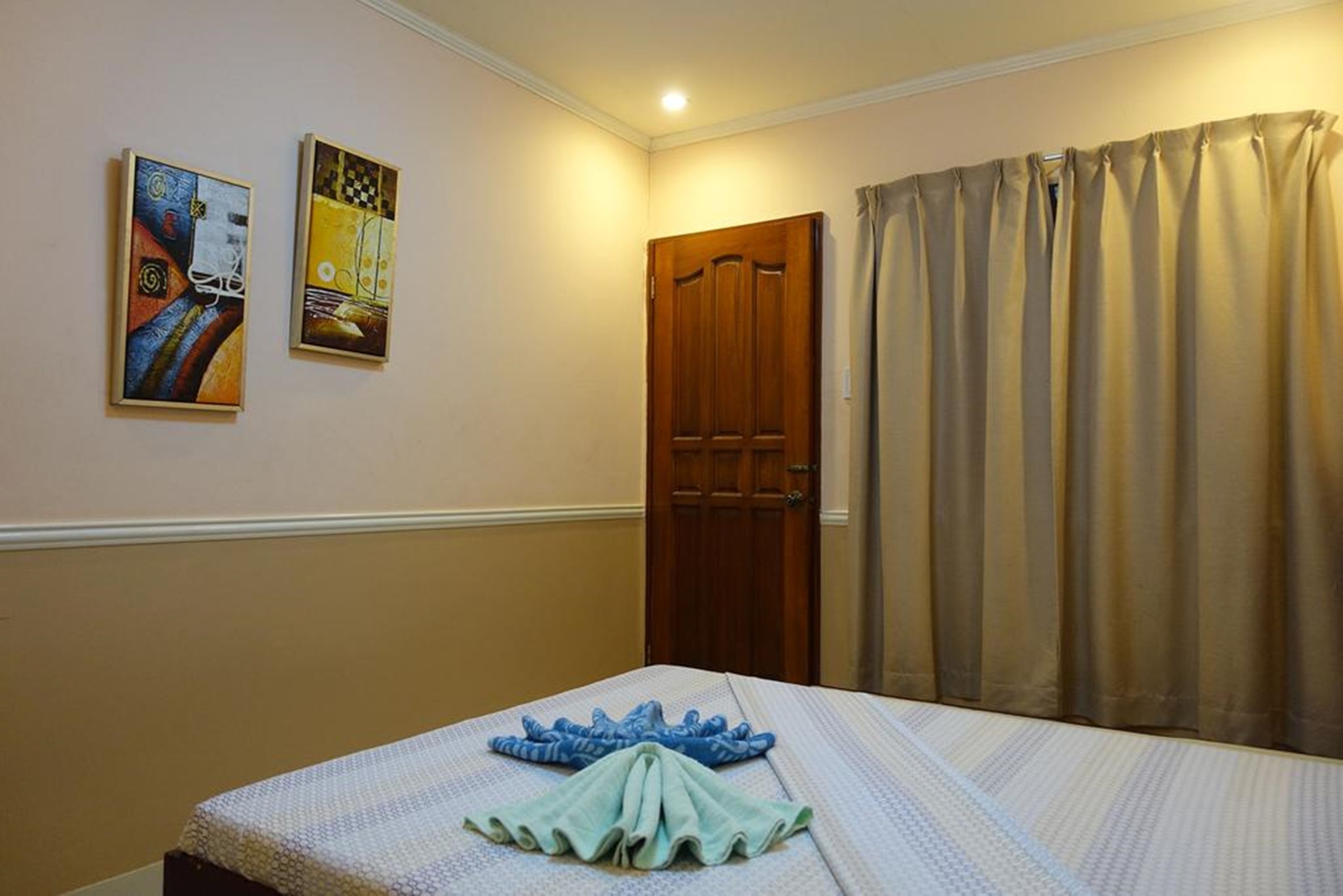 Habitación de hotel con cama, dos cuadros en la pared, una puerta de madera y cortinas color beige.