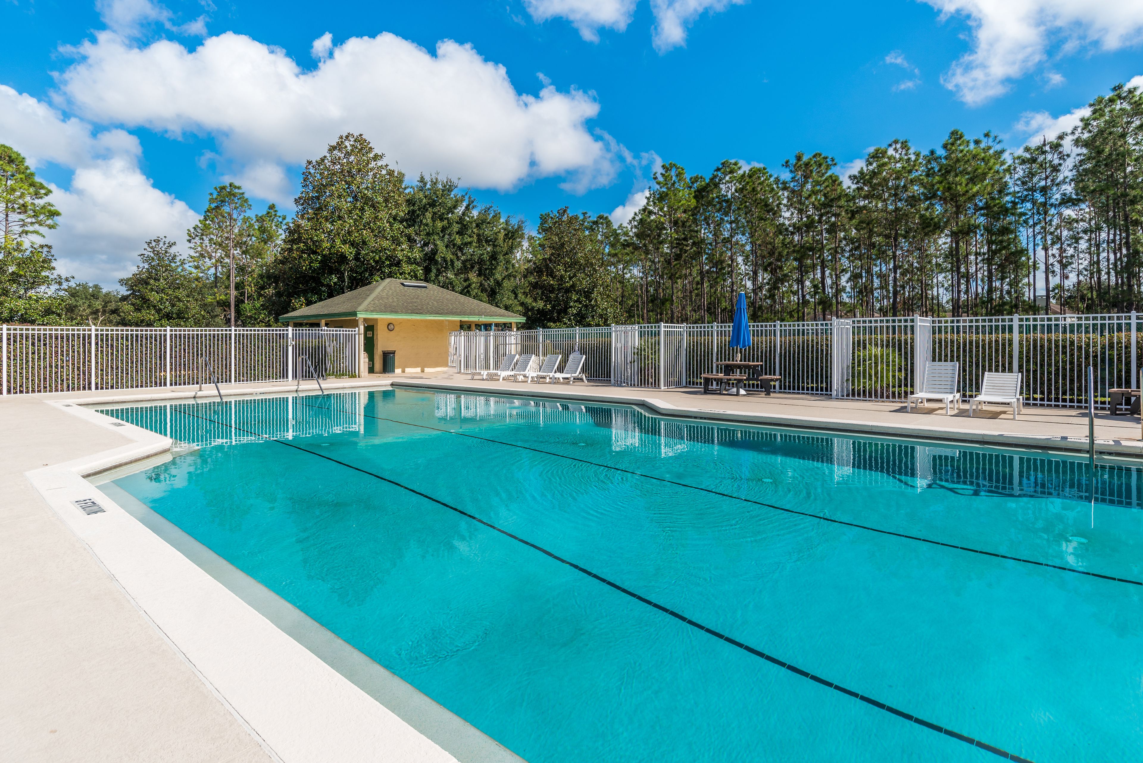 Une piscine entourée d’une clôture, avec des chaises longues et un petit bâtiment en arrière-plan.