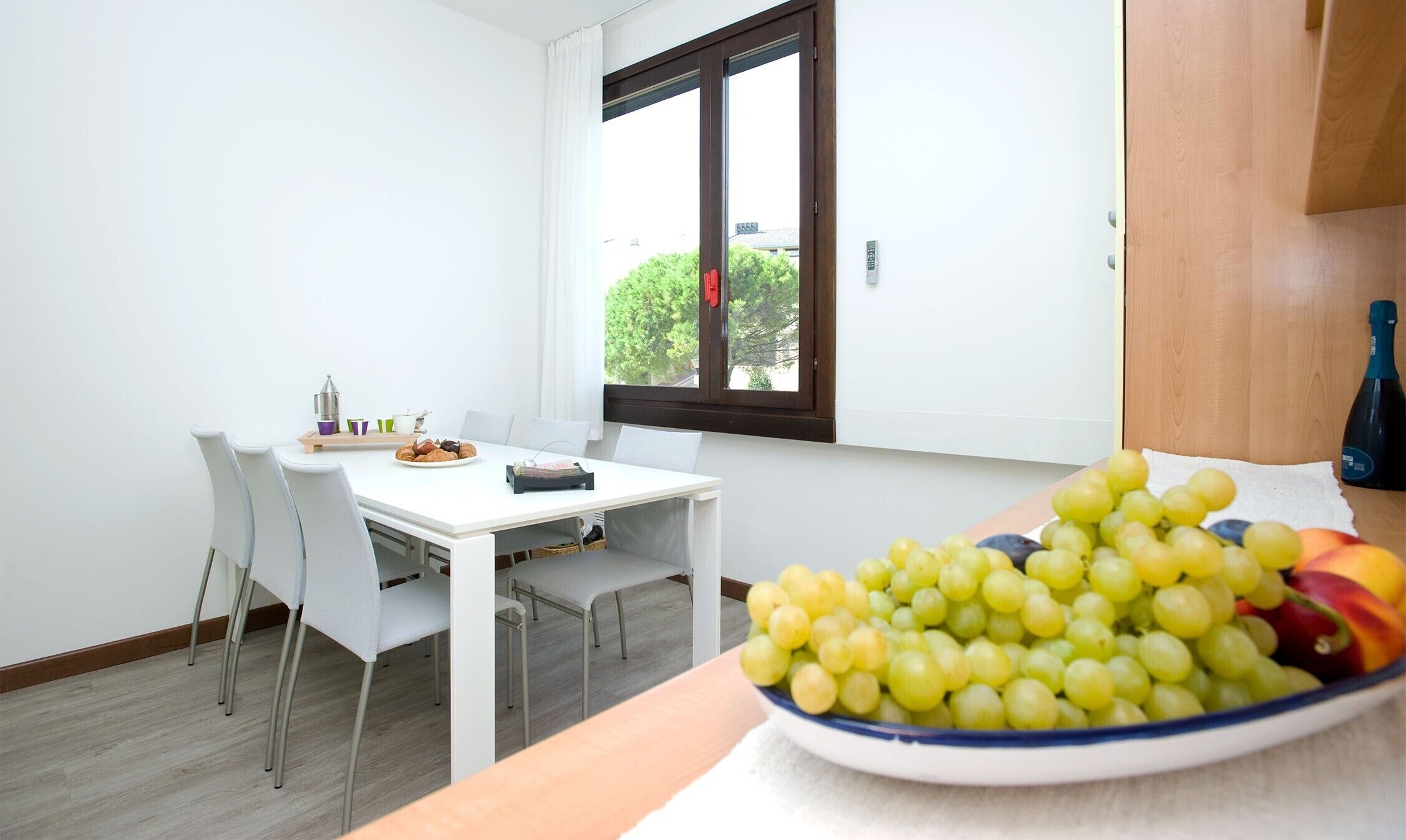 Un comedor con una mesa y sillas blancas, una ventana con vistas a la vegetación y un frutero sobre la mesa.
