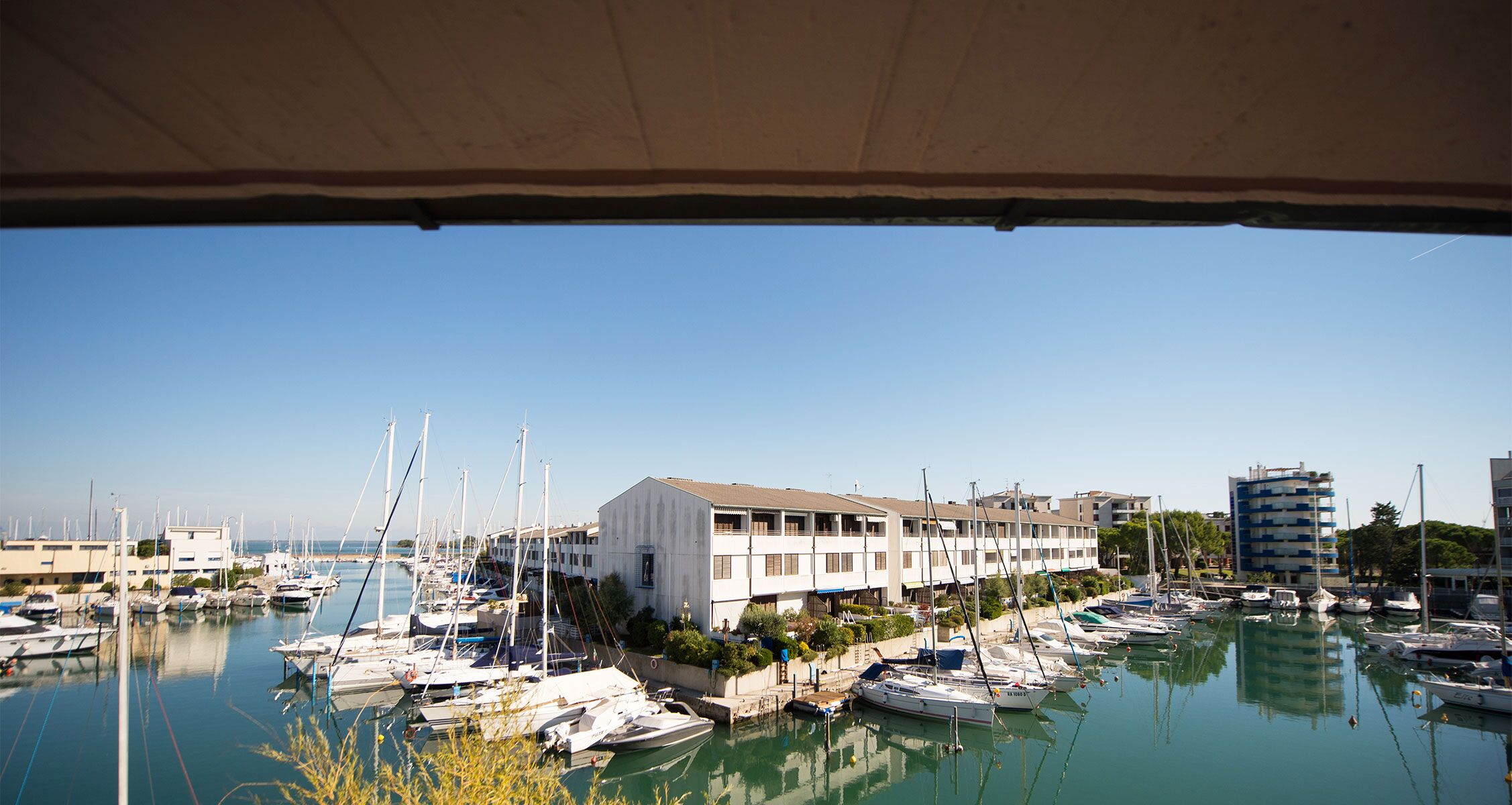 Un puerto deportivo con varias embarcaciones amarradas, un edificio de techo plano y un cielo azul despejado.