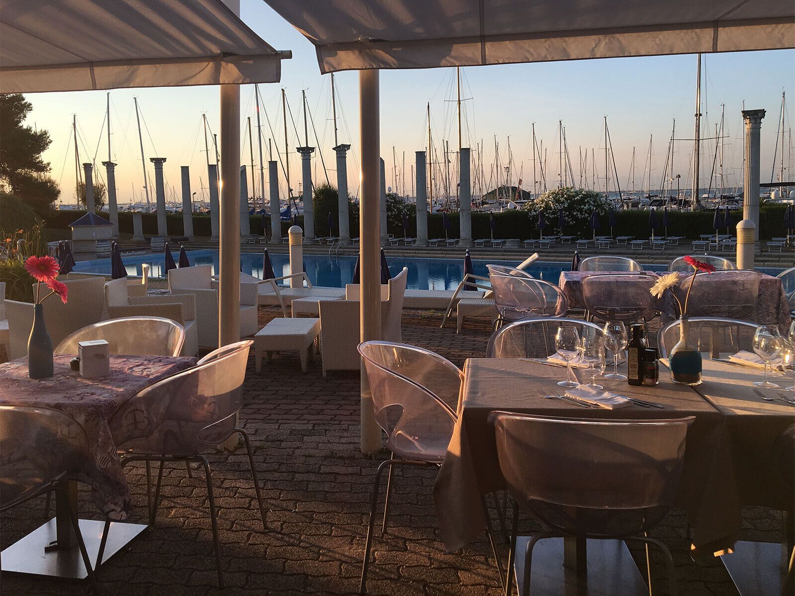Uma área de refeições ao ar livre com mesas e cadeiras, uma piscina e um porto com muitos barcos ao fundo.