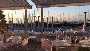 An outdoor dining area with tables and chairs, a pool, and a marina with numerous boats in the background. - Isola Terramare (Lignano Sabbiadoro)