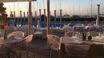 Uma área de refeições ao ar livre com mesas e cadeiras, uma piscina e um porto com muitos barcos ao fundo.