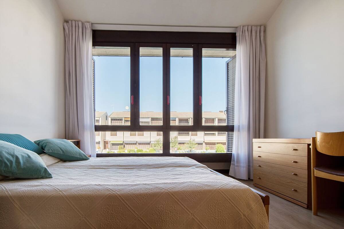 Un dormitorio con una cama, dos almohadas, un aparador de madera y una ventana con cortinas.