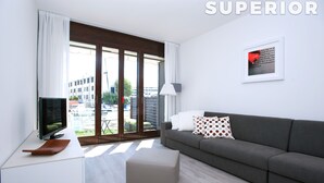 A modern living room with a grey sofa, a TV on a white stand, a wooden side table, and a large window with a view of buildings and greenery. - Isola Terramare (Lignano Sabbiadoro)
