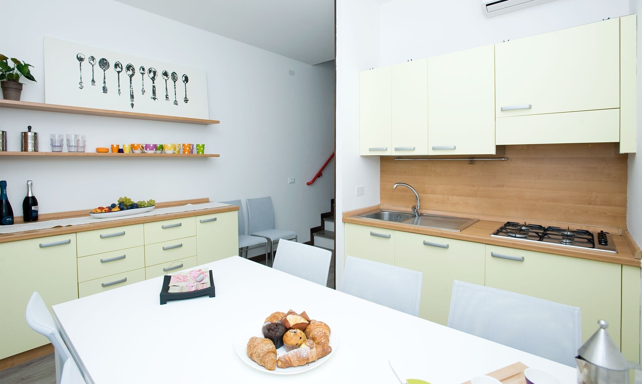 A modern kitchen with light green cabinets, a white countertop, and a stainless steel sink. There is a dining table with chairs and a plate of pastries.