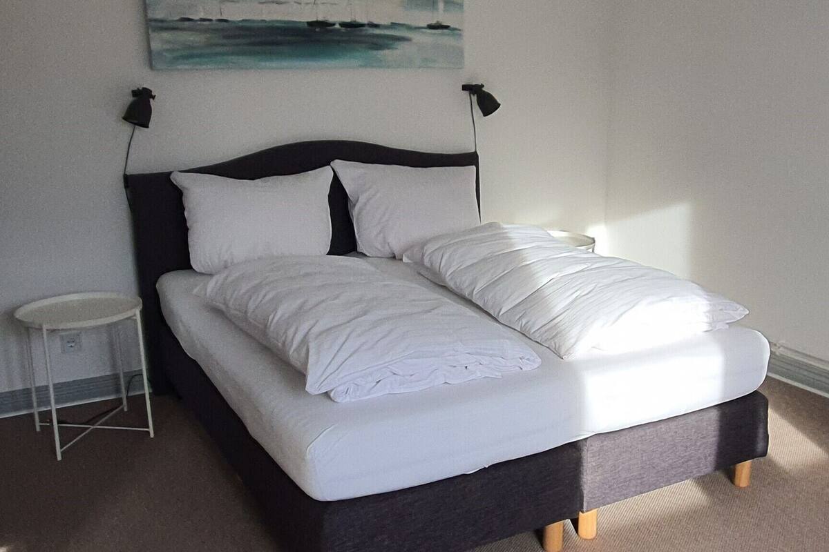 Una cama con ropa de cama blanca, una mesita de noche pequeña y un cuadro en la pared.