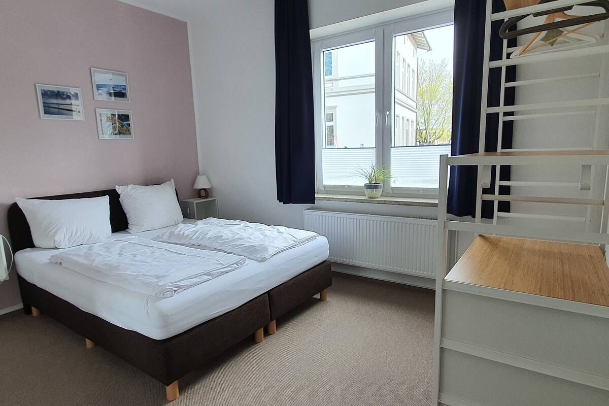 Una cama bien hecha en una habitación con ventana, un radiador y una mesita de noche con lámpara.