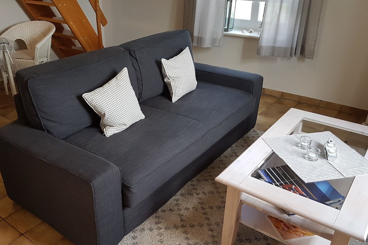 Un sofá gris oscuro con cojines estampados, una mesa de centro blanca con revistas, y una silla de mimbre en una sala de estar con piso de baldosas.
