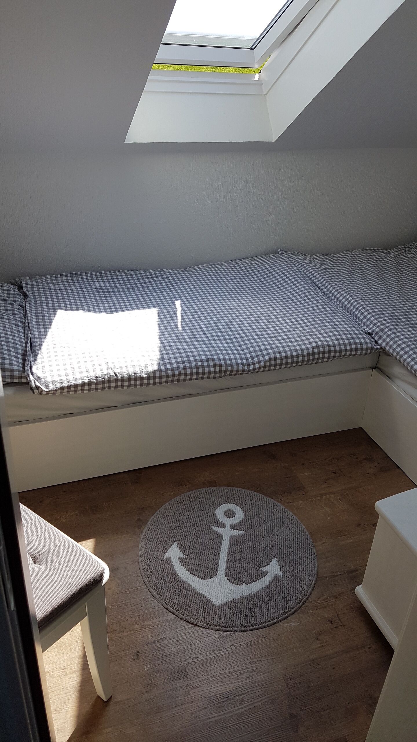 A room with a skylight, a checkered bed, a round rug with an anchor design, and a wooden floor.