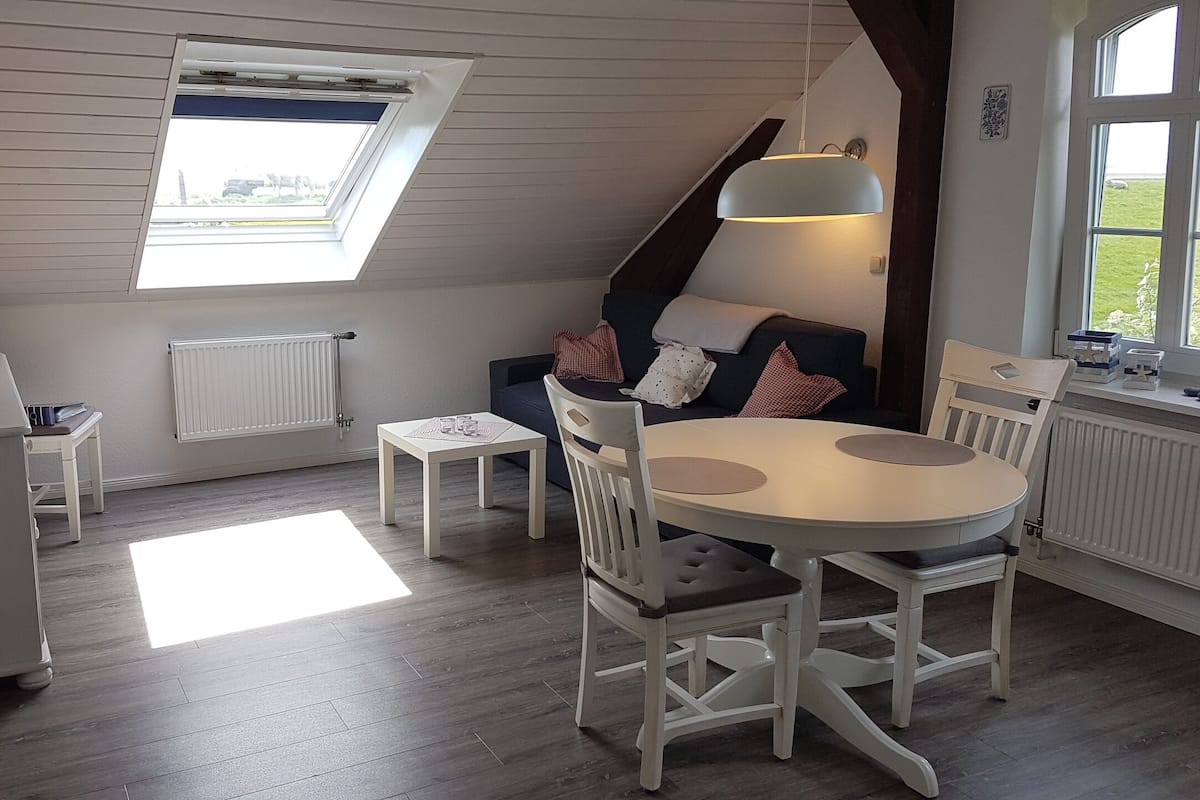 Una habitación acogedora con techo inclinado, un tragaluz, una mesa de comedor con sillas, un sofá con cojines, un radiador y una ventana con vistas al exterior.