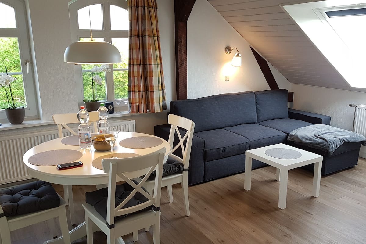 Una habitación acogedora con un sofá de color gris oscuro, dos mesitas de noche blancas, una mesa redonda con sillas y un tragaluz.