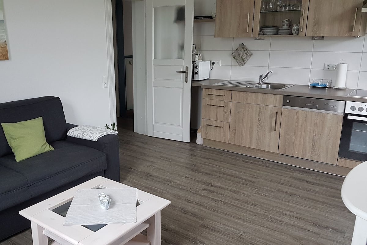 Una sala de estar moderna con un sofá negro, una mesita blanca y un cuadro con barcos en la pared. La cocina tiene armarios de madera, un fregadero y una estufa.
