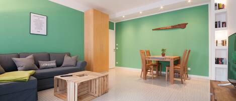 Una sala de estar moderna con una pared verde, un sofá gris, una mesa de centro de madera y un comedor con una mesa y sillas de madera.