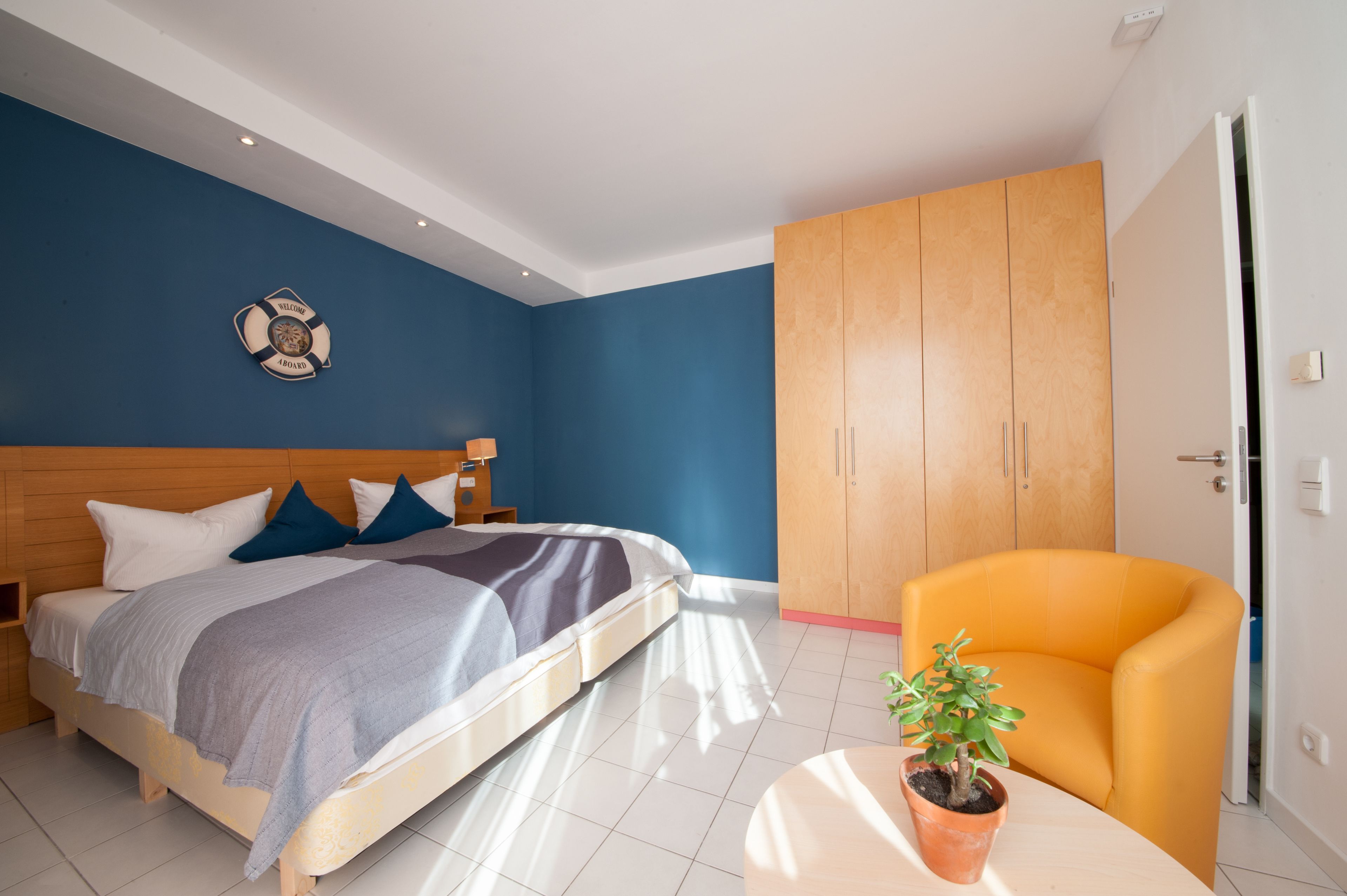 Um quarto com uma cama, uma mesa de cabeceira, um guarda-roupa de madeira, uma poltrona amarela e um vaso com planta.