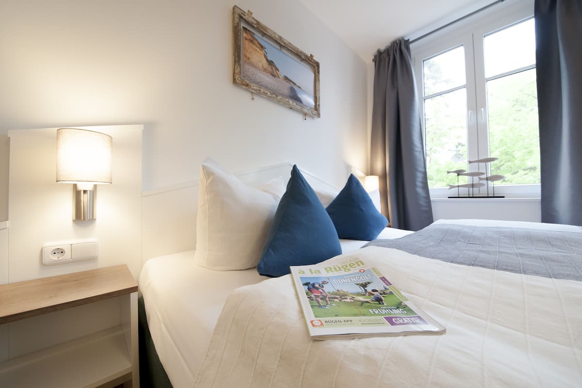 A hotel room with a bed, pillows, a magazine, a lamp, a painting, and a window with curtains.