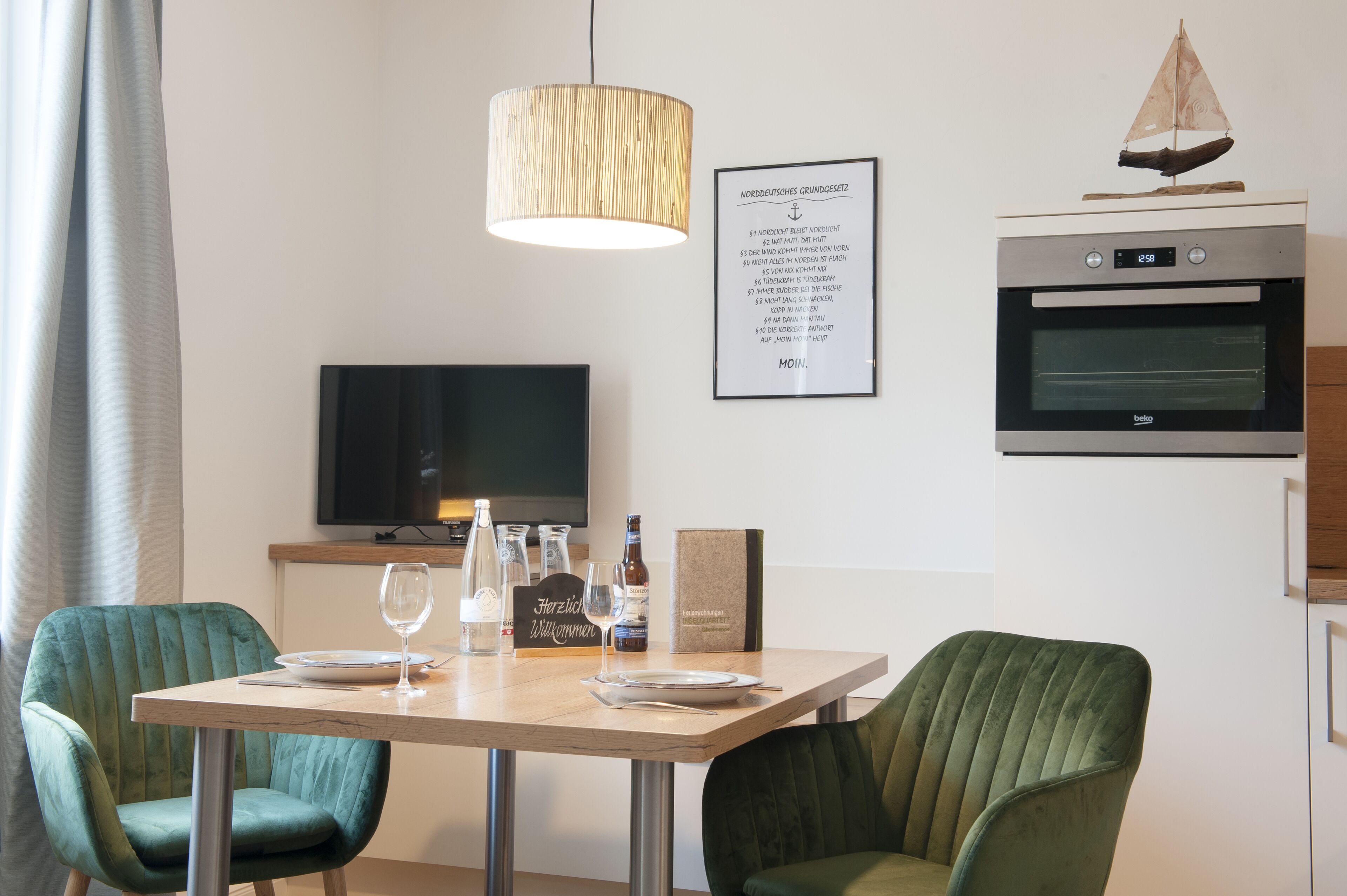Et moderne spiseområde med et træbord, to grønne polstrede stole, en hængende lampe, et fjernsyn og en indramet menu på væggen.