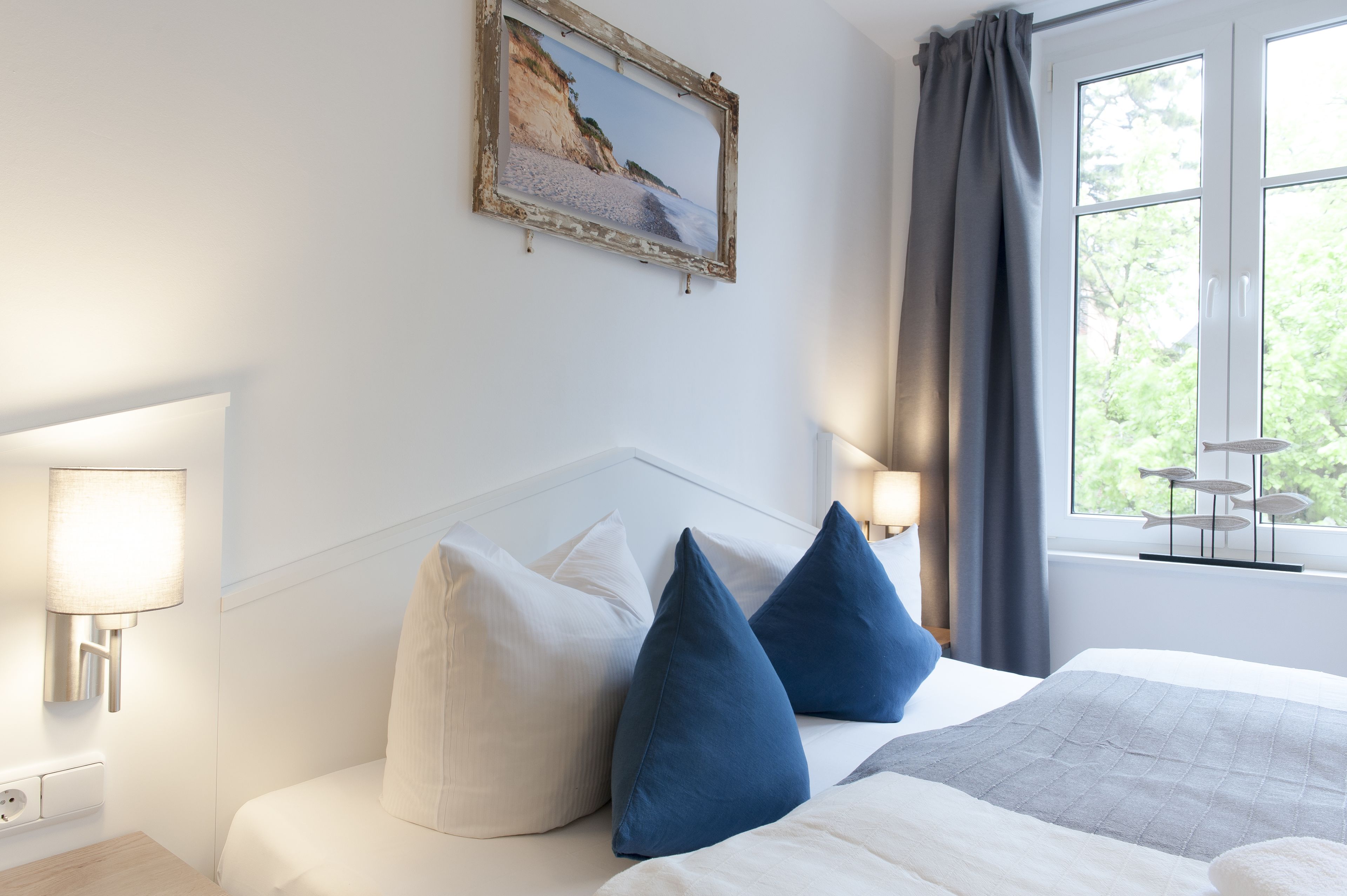 Una habitación de hotel con cama, almohadas blancas y azules, un cuadro de una playa, una lámpara y una ventana con cortinas.