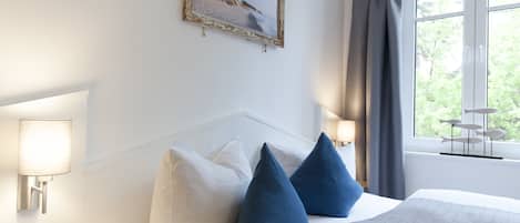Una habitación de hotel con cama, almohadas blancas y azules, un cuadro de una playa, una lámpara y una ventana con cortinas.