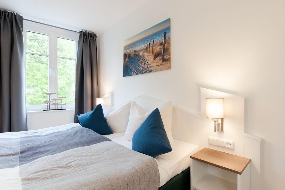 A hotel room with a bed, a bedside table, a nightstand, a wall-mounted lamp, and a window with curtains.