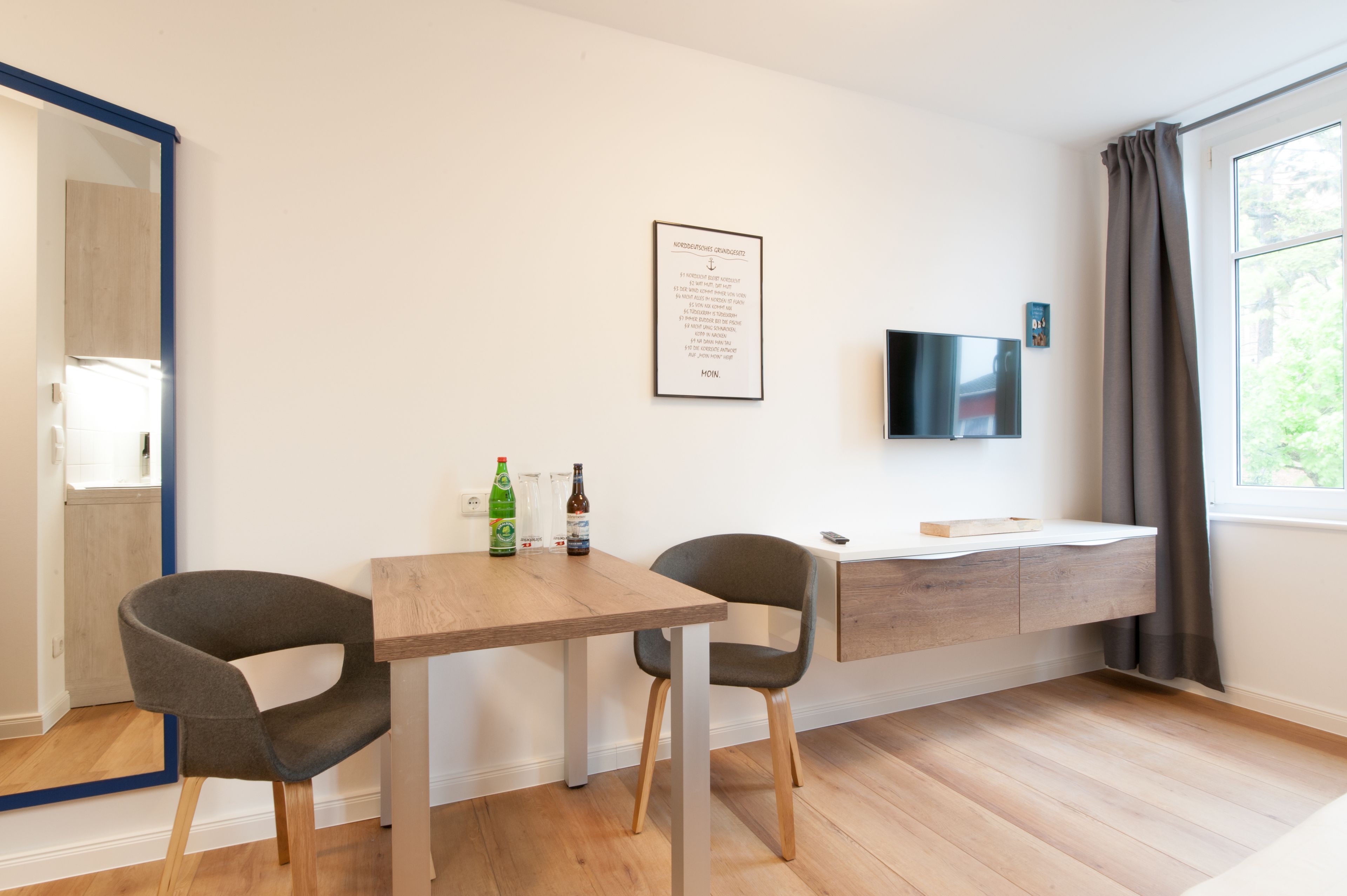 Una habitación moderna con una mesa de madera, dos sillas, un televisor montado y un ventanal con cortinas.