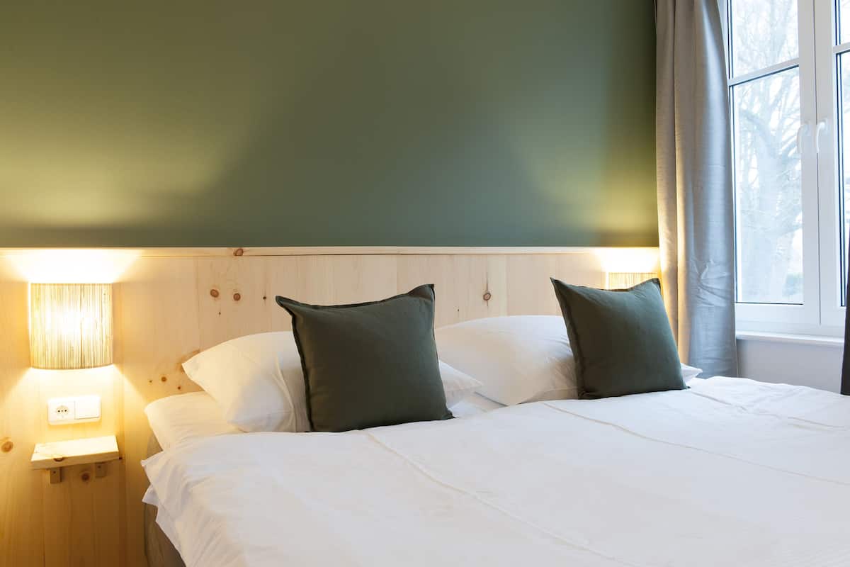 A neatly made bed with white linens and two dark green pillows, a wooden headboard, and a bedside lamp on a nightstand.
