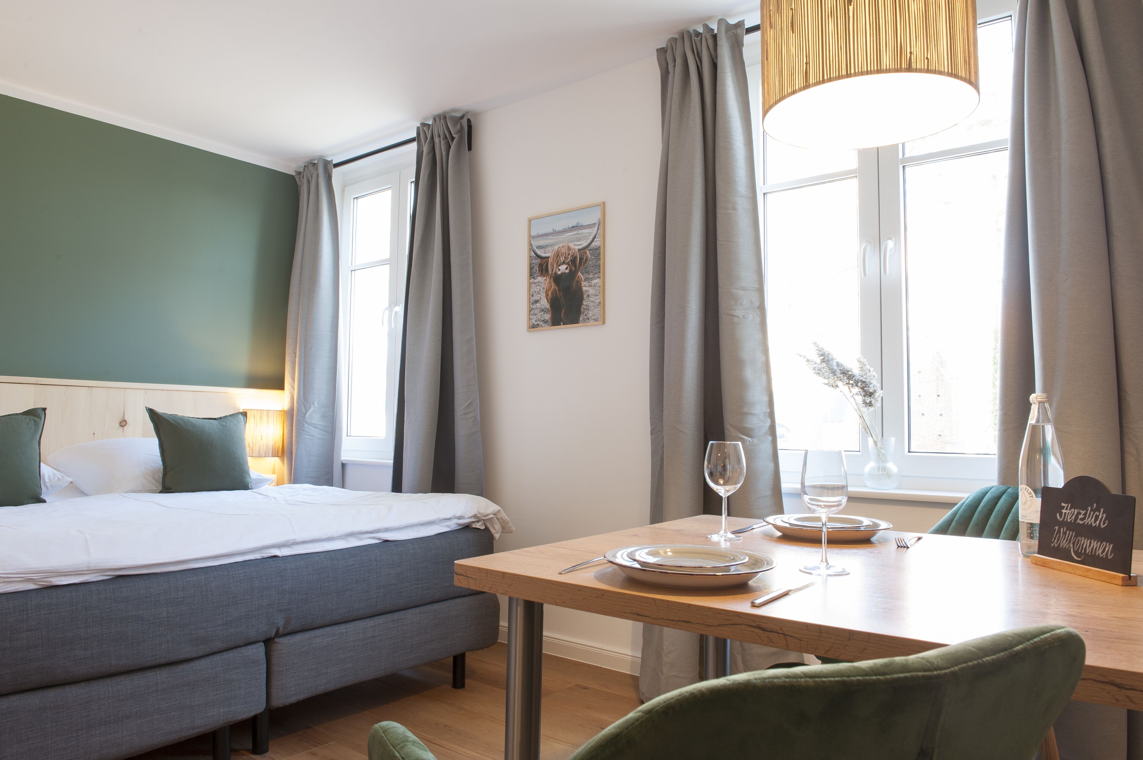 A hotel room with a bed, a dining table with a glass of water, a bottle, and a sign that says "Herzlich Willkommen".