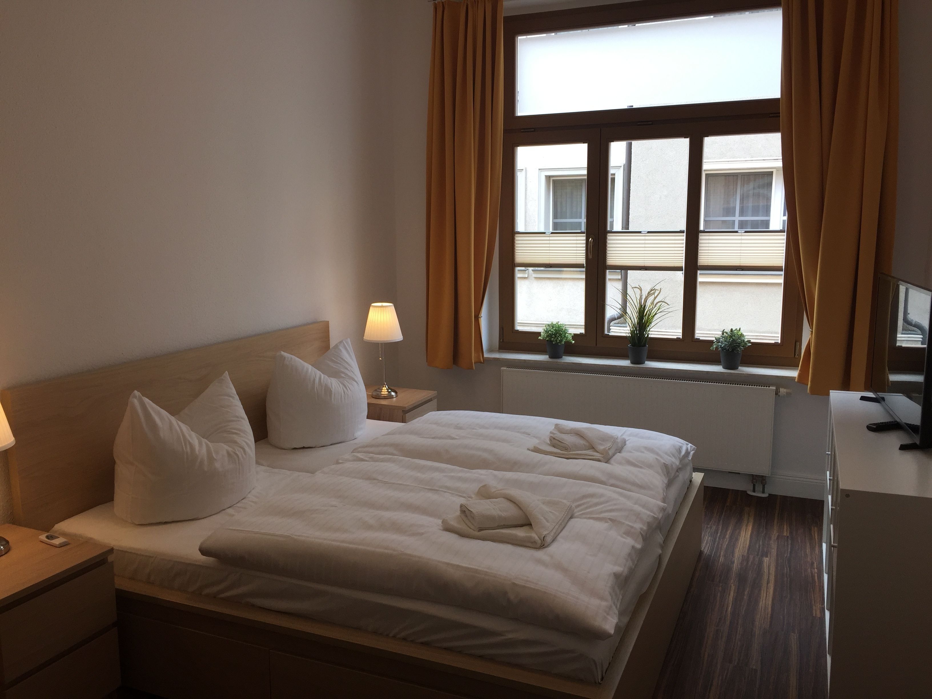 A hotel room with a bed, bedside tables, a desk, a TV, and a window with curtains.