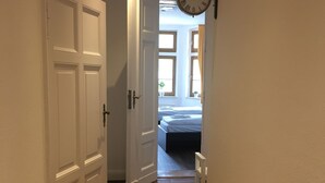 Een smalle gang met houten vloer, witte wanden en een witte deur aan de linkerkant. Een klok hangt aan de rechtermuur.