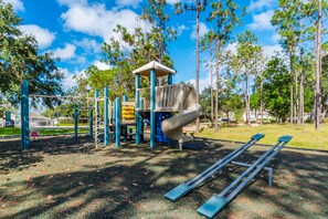 Explore a playground with a slide, swings, and climbing elements set amidst a lush green area surrounded by trees.
