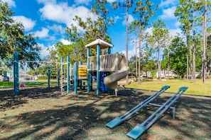 Explore a playground with a slide, swings, and climbing elements set amidst a lush green area surrounded by trees.