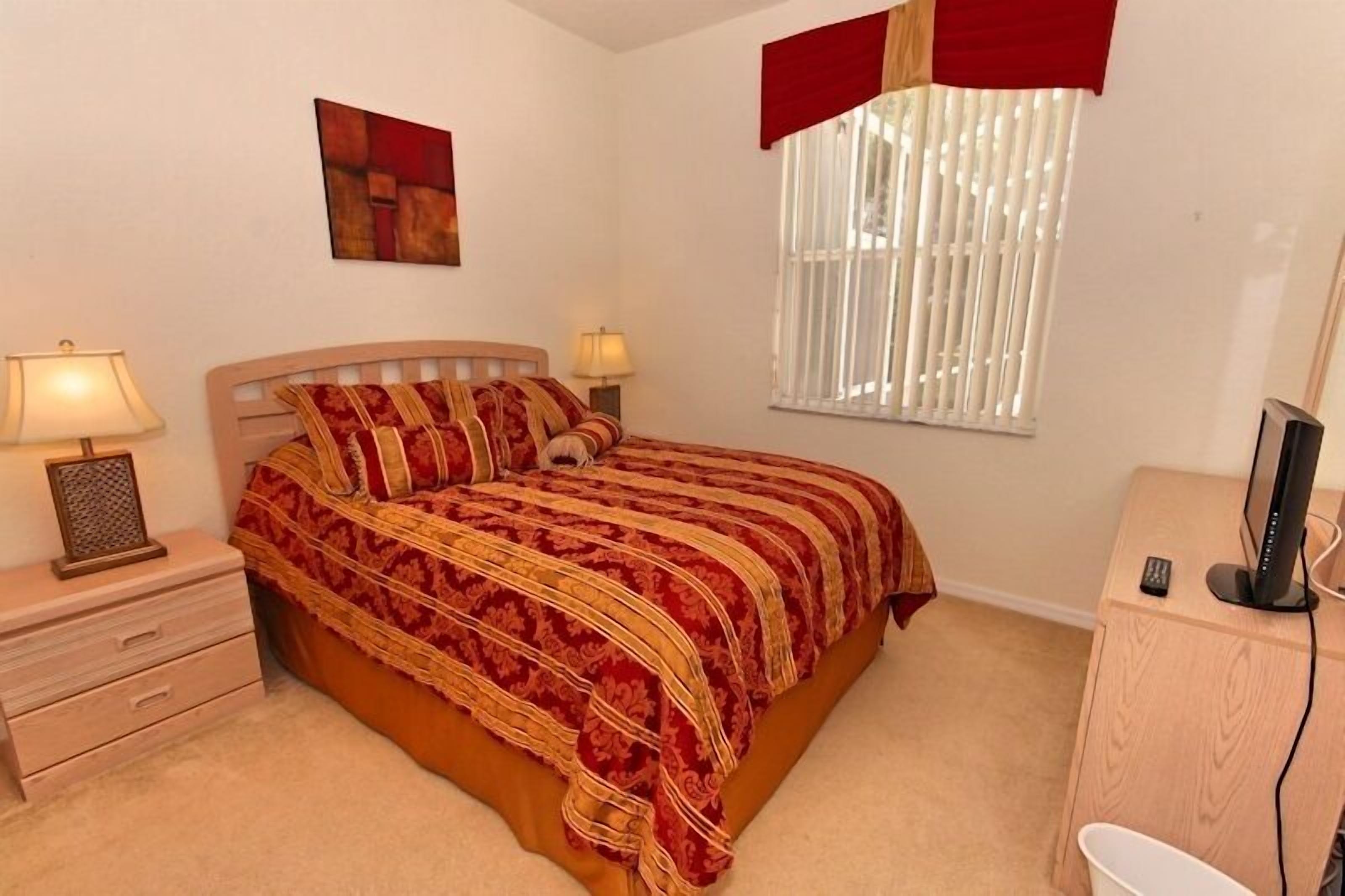 A bedroom with a bed, two bedside lamps, a TV, a desk, and a window with blinds.