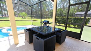 Un patio al aire libre con una mesa de vidrio y sillas de mimbre negro, con vistas a una piscina y un césped bien cuidado.