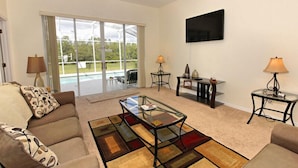 A spacious living room with a glass coffee table, a sofa, a TV mounted on the wall, and a view of the outdoors through large sliding doors. - Ov3123 - Highlands Reserve - 4 Bed 3 Baths Villa (Davenport)
