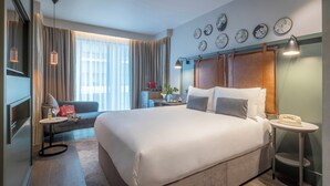 Une chambre d’hôtel avec un grand lit, un bureau avec une lampe, une chaise, une petite table avec un téléphone, une fenêtre avec des rideaux et des éléments de décoration murale.