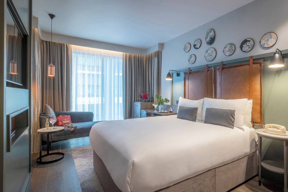 A hotel room with a large bed, a desk with a lamp, a chair, a small table with a phone, a window with curtains, and wall decor.