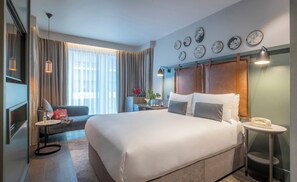 Habitación de hotel con una cama grande, un escritorio con lámpara, una silla, una mesita con teléfono, una ventana con cortinas y decoración en la pared.
