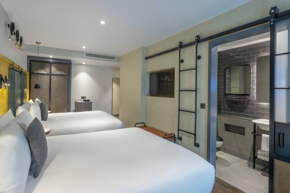A hotel room with two beds, a small desk, a mirror, and a bathroom visible through an open door.