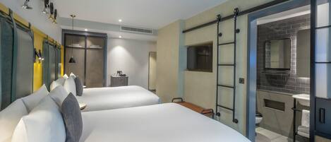 Une chambre d’hôtel avec deux lits, un petit bureau, un miroir et une salle de bain visible à travers une porte ouverte.
