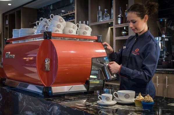 A barista preparing coffee at a commercial espresso machine, with cups and a muffin on the counter. - Clayton Hotel City of London, Tower Bridge (London)