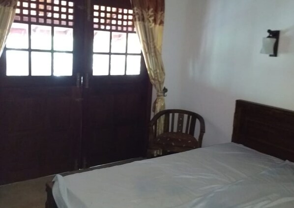 Quarto com cama, cadeira de madeira, janela com cortinas e luminária na parede.